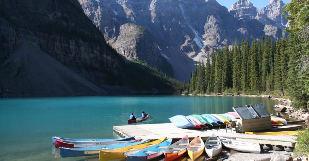 Two people on a canoe with a line of canoes tied to a dock in front of them and mountains and trees in the background.