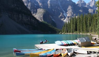 Two people on a canoe with a line of canoes tied to a dock in front of them and mountains and trees in the background.